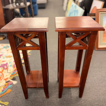 A pair of tall slim wooden side tables featuring square tops, lower display shelves, and cross-brace side details.