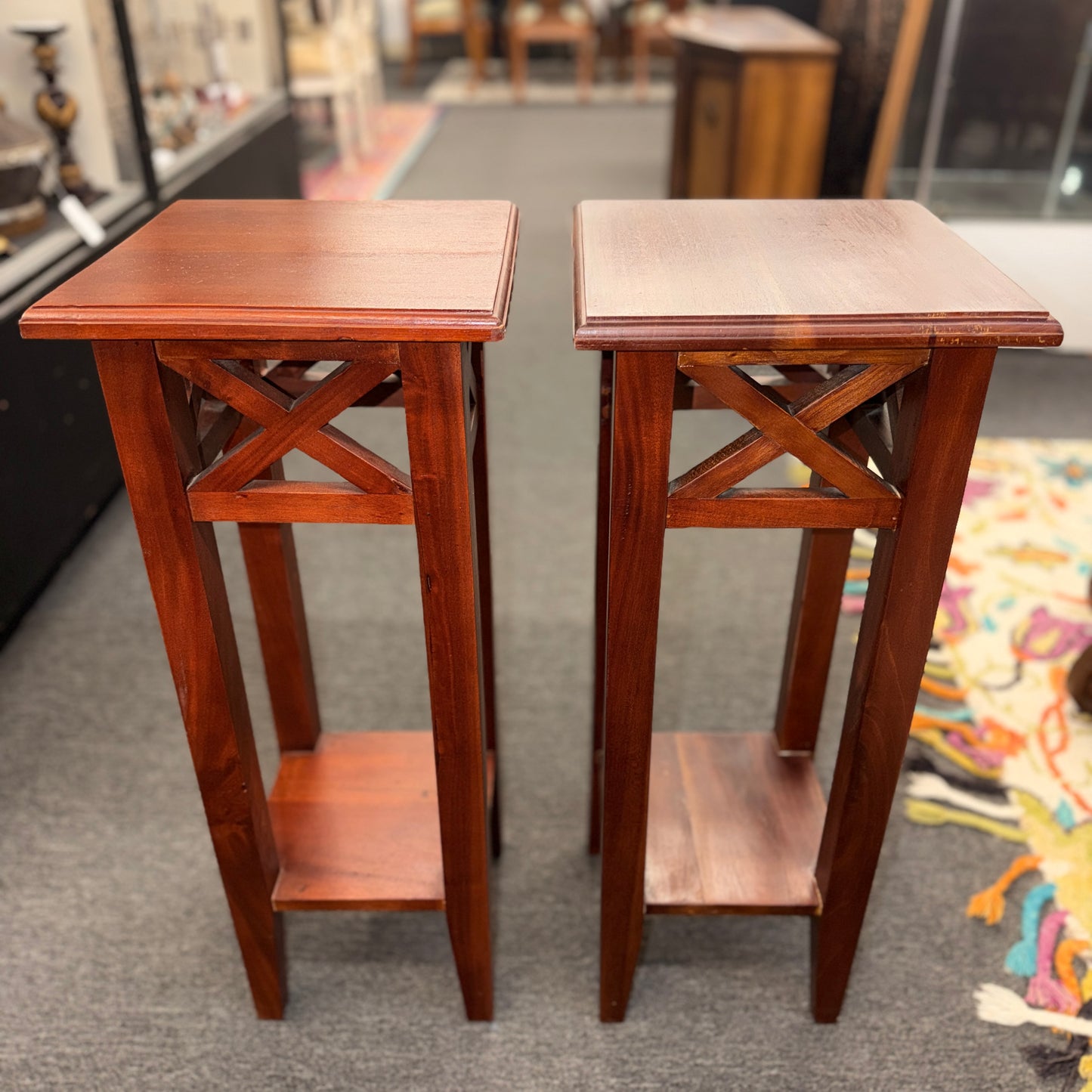 A pair of tall slim wooden side tables featuring square tops, lower display shelves, and cross-brace side details.