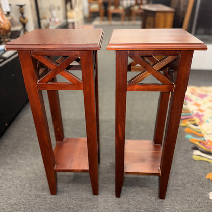 A pair of tall slim wooden side tables featuring square tops, lower display shelves, and cross-brace side details.