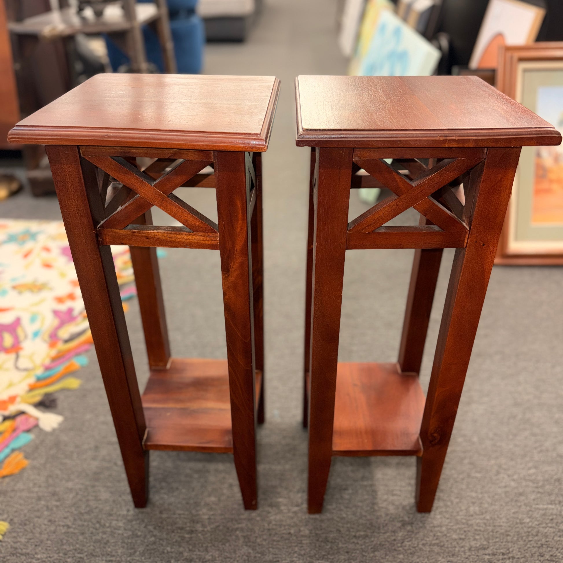 A pair of tall slim wooden side tables featuring square tops, lower display shelves, and cross-brace side details.