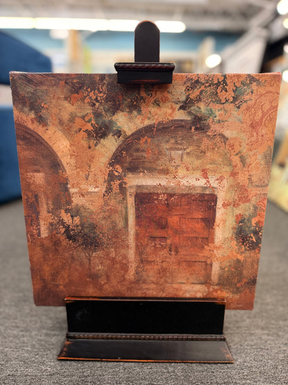 A textured square canvas painting of weathered Mediterranean arches mounted on a black ornamental display stand in excellent condition.