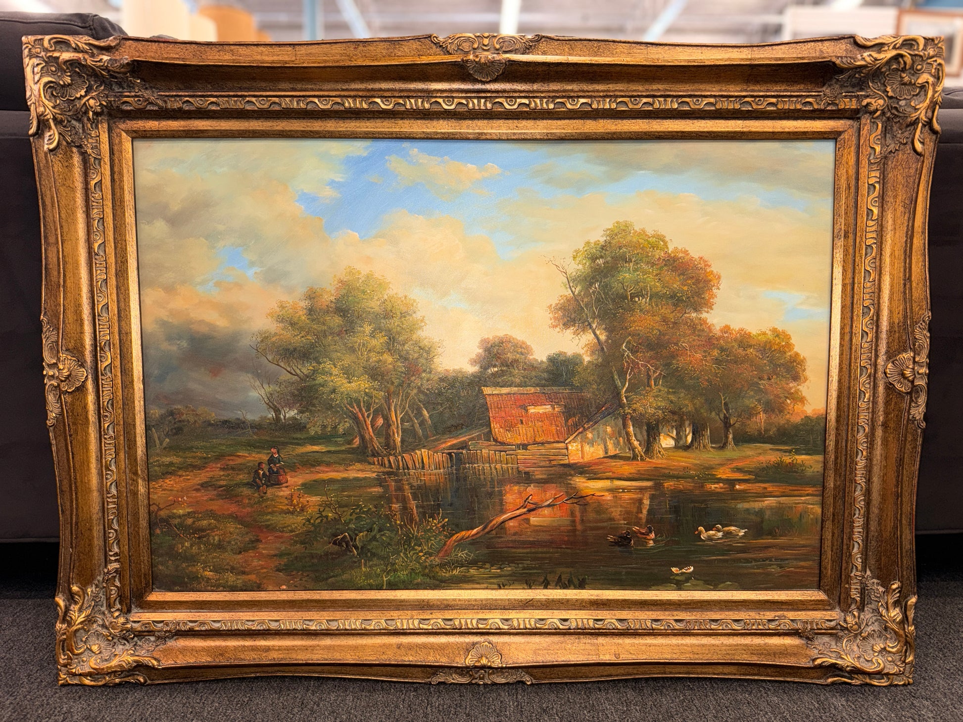 A traditional pastoral landscape painting depicting a lakeside cabin, autumn trees, and ducks in an ornate carved metallic-gold frame in excellent condition.