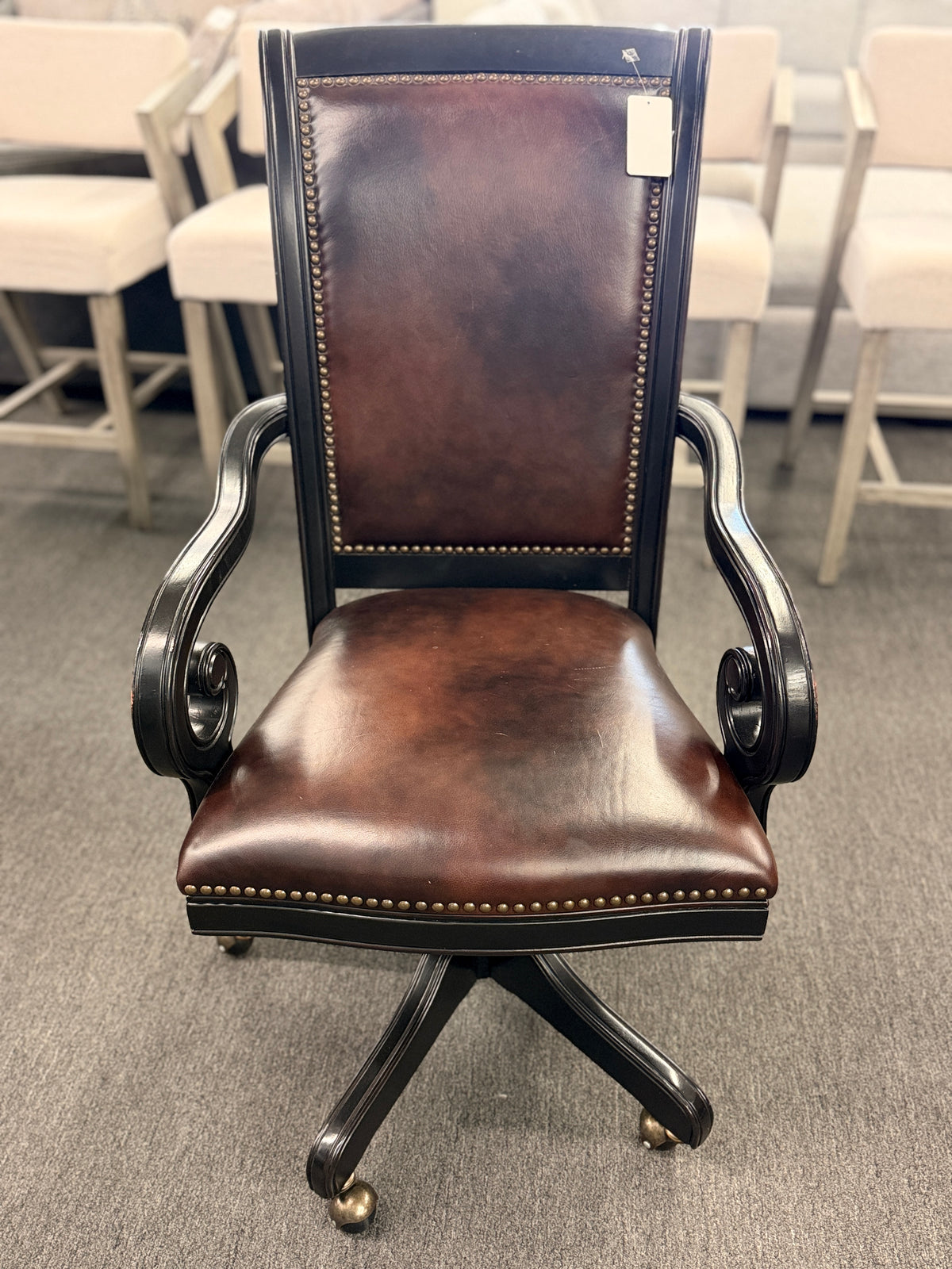 An executive traditional swivel desk chair featuring rich brown faux-leather upholstery with brass nailhead trim, scrolled wooden armrests, and a rolling caster base in good condition.