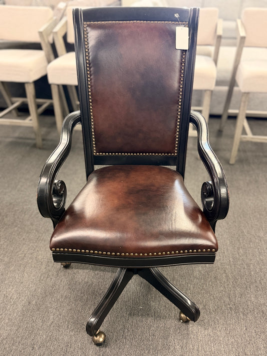 An executive traditional swivel desk chair featuring rich brown faux-leather upholstery with brass nailhead trim, scrolled wooden armrests, and a rolling caster base in good condition.