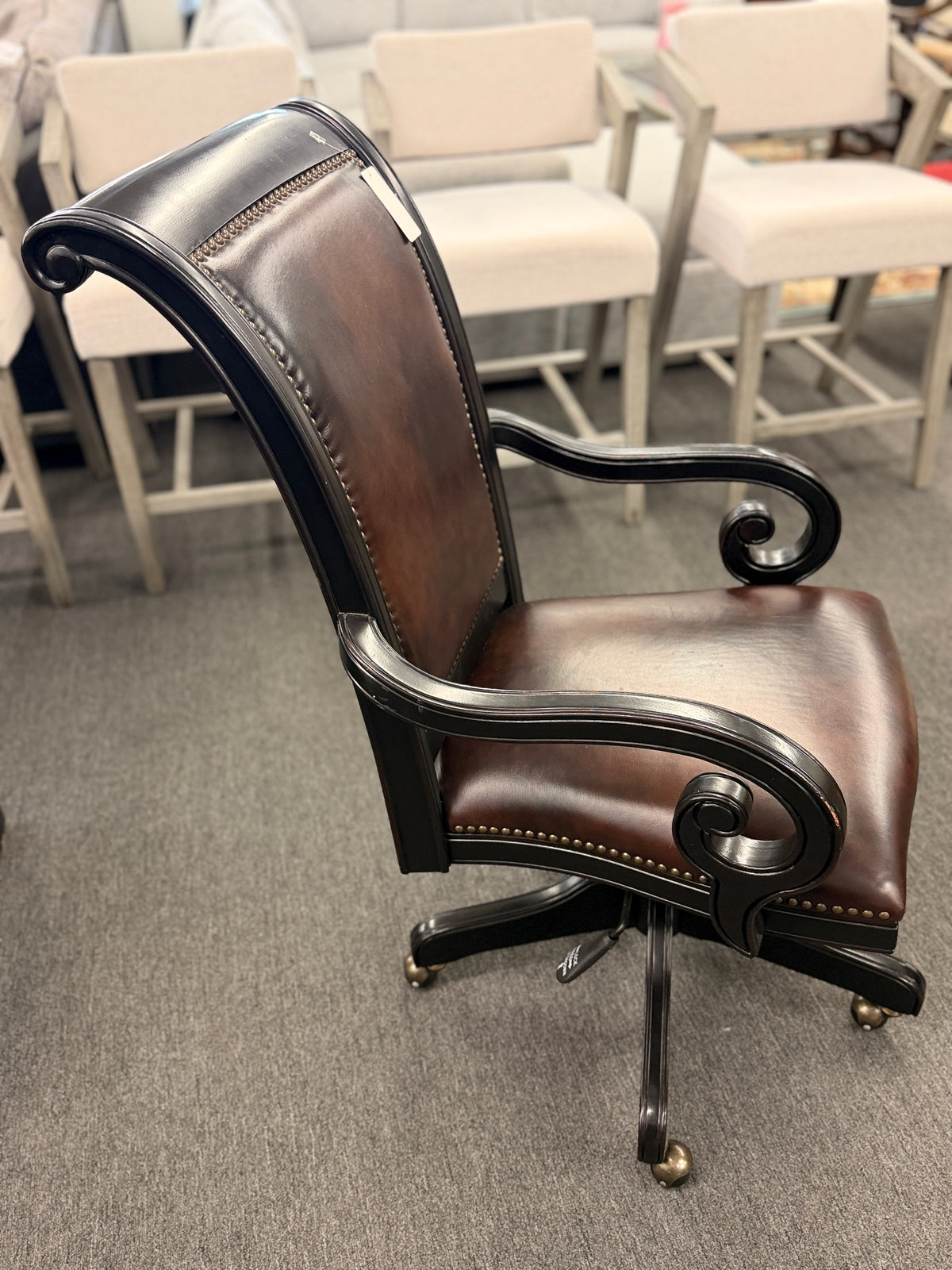 An executive traditional swivel desk chair featuring rich brown faux-leather upholstery with brass nailhead trim, scrolled wooden armrests, and a rolling caster base in good condition.