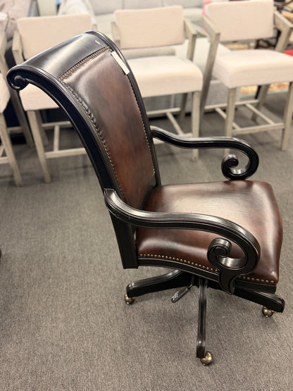 An executive traditional swivel desk chair featuring rich brown faux-leather upholstery with brass nailhead trim, scrolled wooden armrests, and a rolling caster base in good condition.
