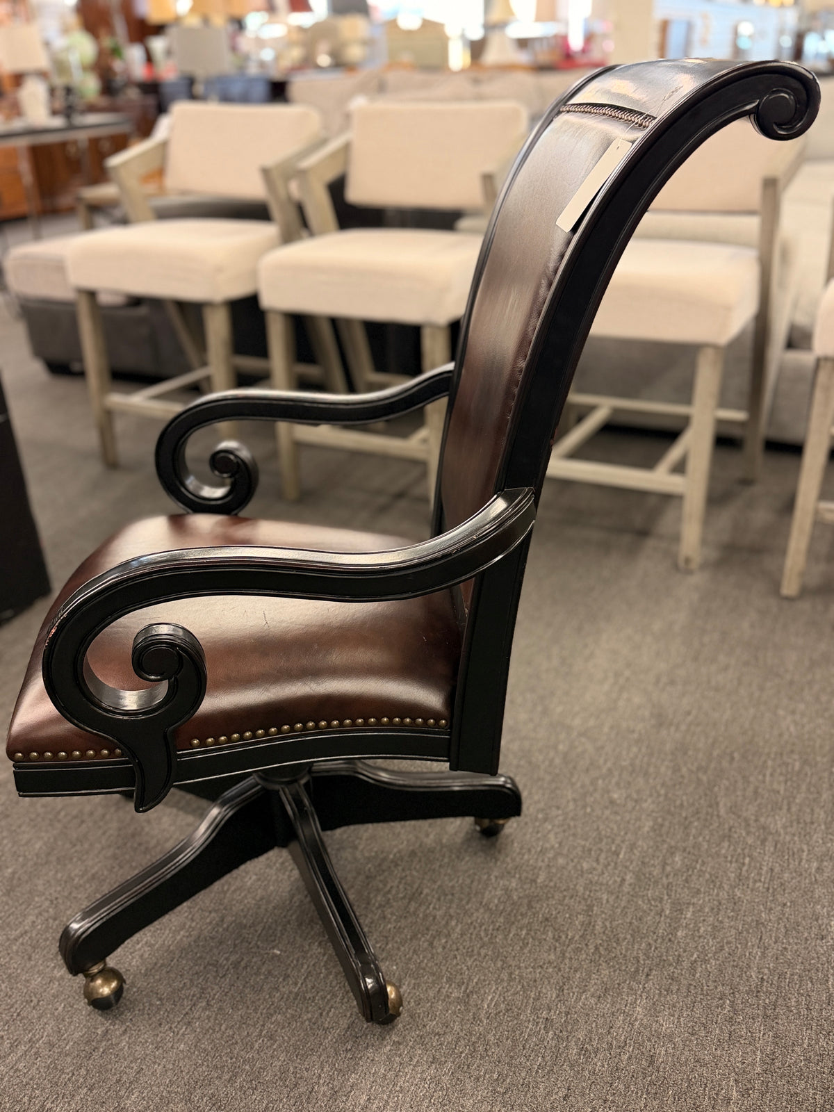 An executive traditional swivel desk chair featuring rich brown faux-leather upholstery with brass nailhead trim, scrolled wooden armrests, and a rolling caster base in good condition.