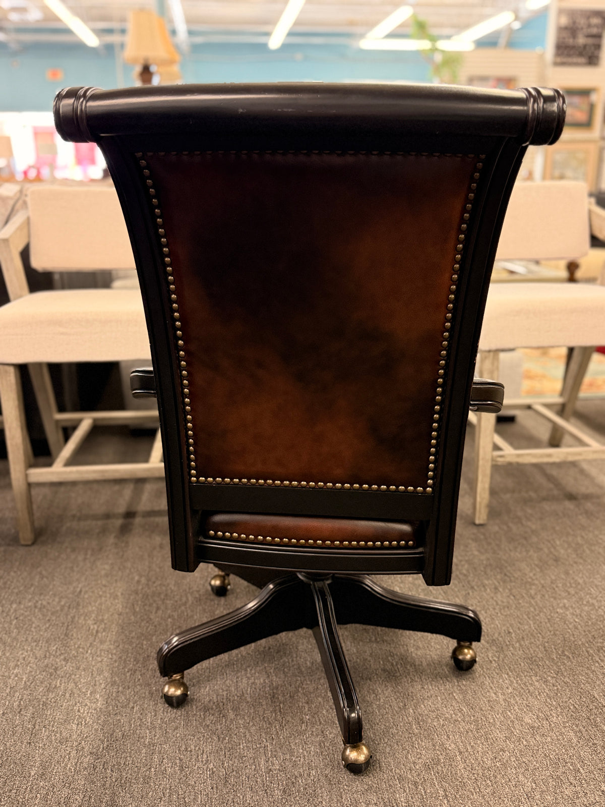An executive traditional swivel desk chair featuring rich brown faux-leather upholstery with brass nailhead trim, scrolled wooden armrests, and a rolling caster base in good condition.