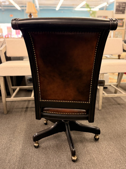 An executive traditional swivel desk chair featuring rich brown faux-leather upholstery with brass nailhead trim, scrolled wooden armrests, and a rolling caster base in good condition.