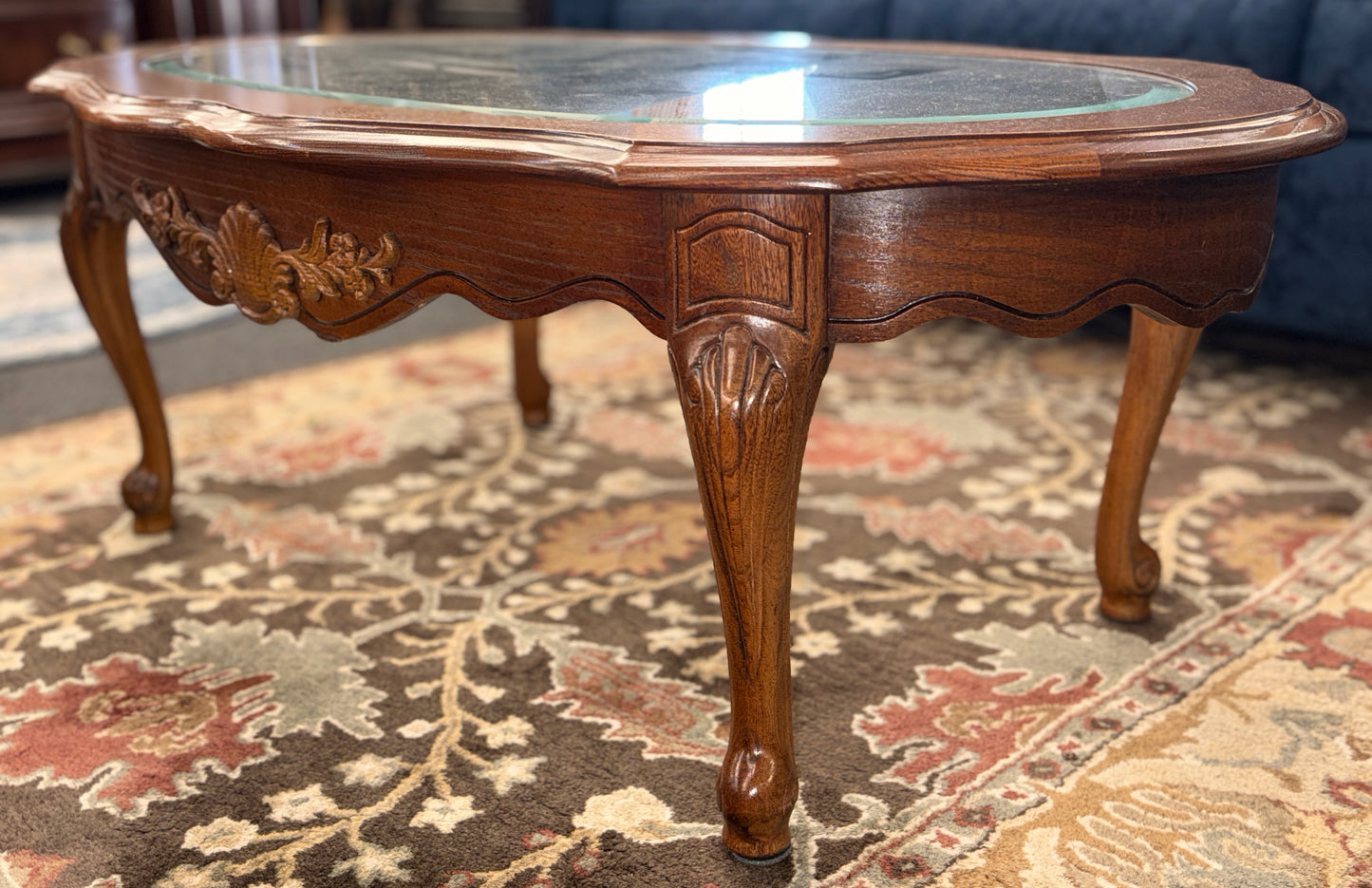 A classic oval coffee table featuring an inset clear glass top, hand-carved shell and floral details on the apron, and sweeping cabriole legs.