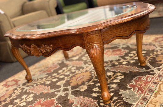A classic oval coffee table featuring an inset clear glass top, hand-carved shell and floral details on the apron, and sweeping cabriole legs.