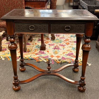 Traditional carved wood console table featuring two front drawers, turned carved legs, an embossed tabletop with nailhead trim, and a curved cross-stretcher with a decorative center finial.