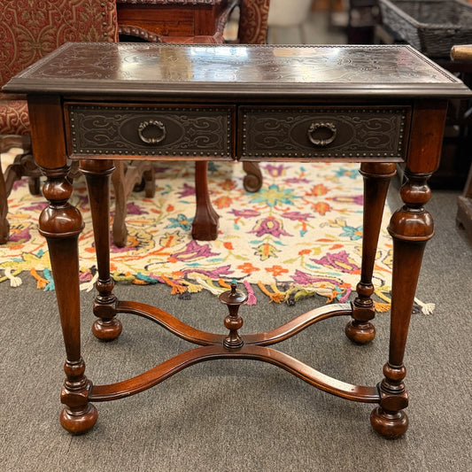 Traditional carved wood console table featuring two front drawers, turned carved legs, an embossed tabletop with nailhead trim, and a curved cross-stretcher with a decorative center finial.