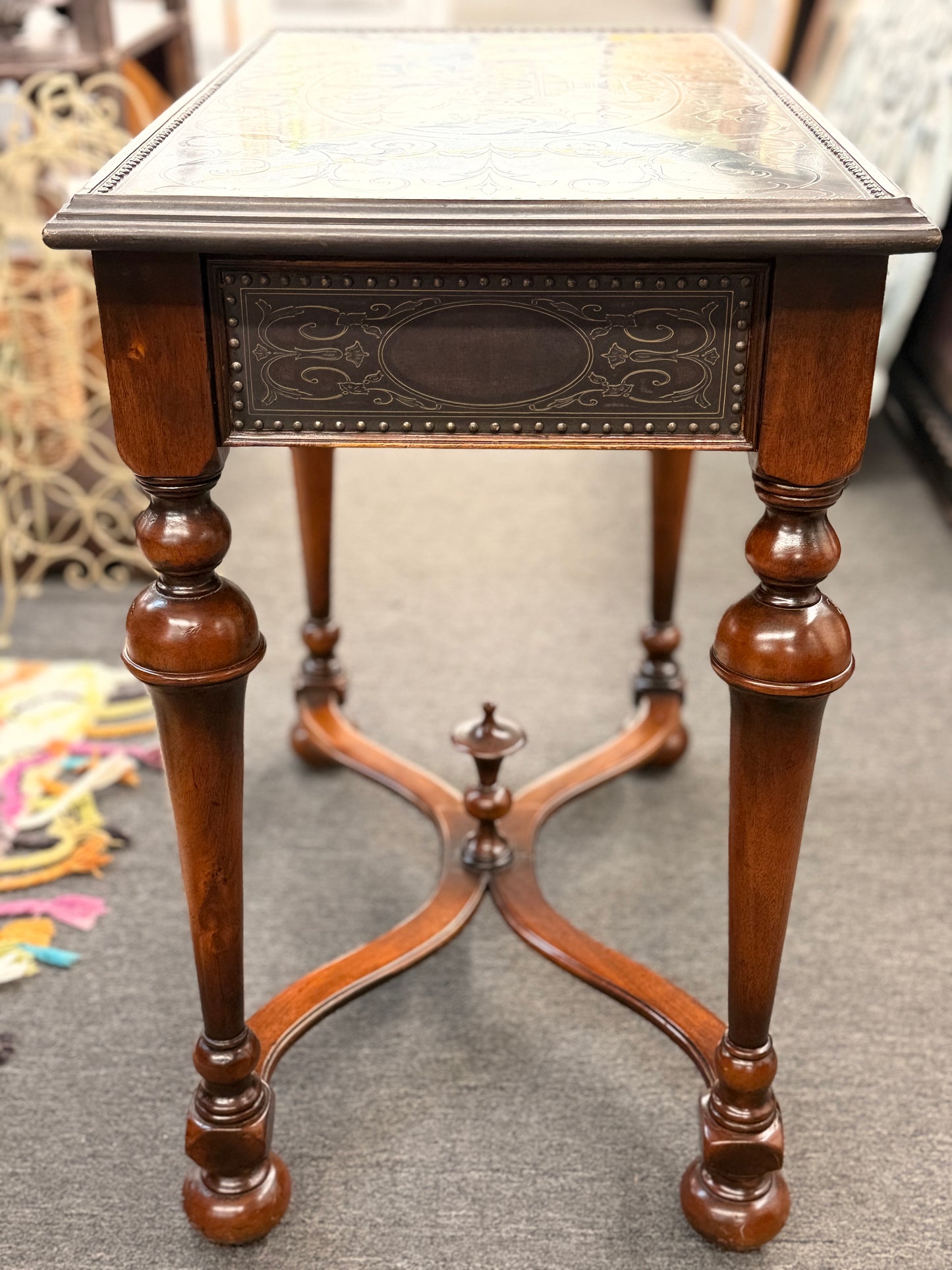 Traditional carved wood console table featuring two front drawers, turned carved legs, an embossed tabletop with nailhead trim, and a curved cross-stretcher with a decorative center finial.