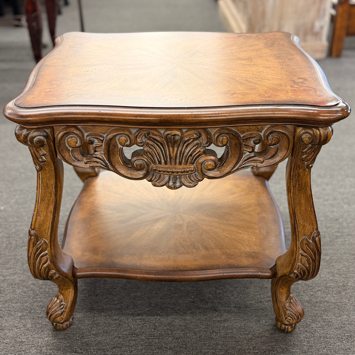 A traditional carved wood end table featuring ornate acanthus leaf aprons, cabriole legs, a sunburst wood veneer top, and a bottom display shelf in good condition.