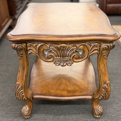 A traditional carved wood end table featuring ornate acanthus leaf aprons, cabriole legs, a sunburst wood veneer top, and a bottom display shelf in good condition.