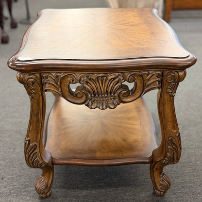 A traditional carved wood end table featuring ornate acanthus leaf aprons, cabriole legs, a sunburst wood veneer top, and a bottom display shelf in good condition.