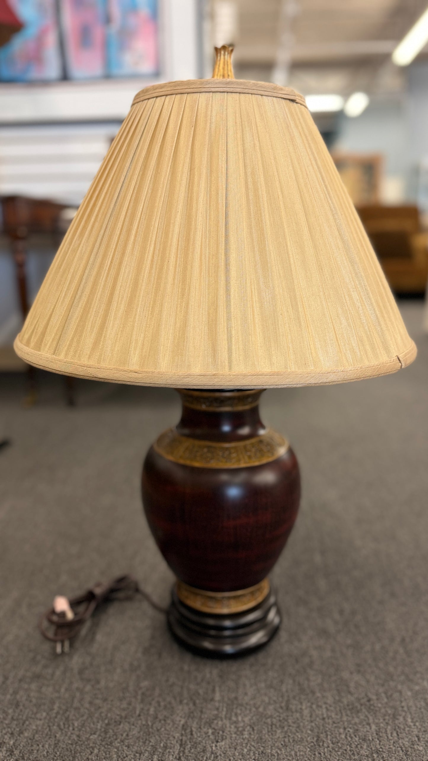 A traditional-style table lamp featuring an urn-shaped ceramic base with decorative gold-toned bands at the neck and base, topped with a pleated flared fabric lampshade.