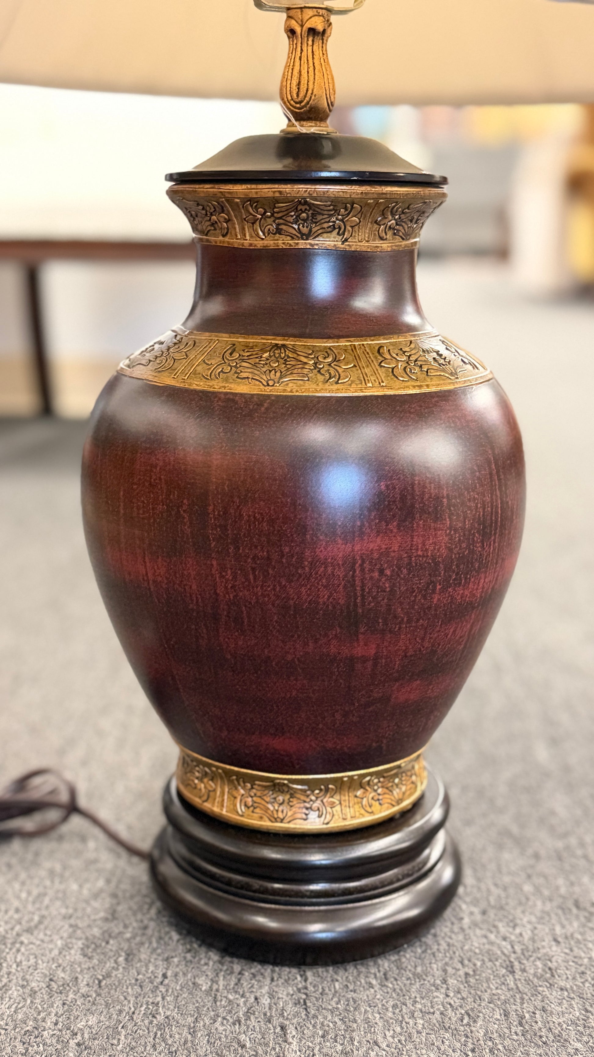 A traditional-style table lamp featuring an urn-shaped ceramic base with decorative gold-toned bands at the neck and base, topped with a pleated flared fabric lampshade.
