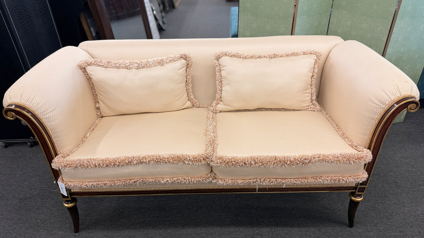 An elegant traditional cream upholstered settee loveseat featuring rolled arms, fringe trim along seat cushions, matching pillows, and fluted wooden legs.