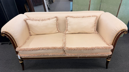 An elegant traditional cream upholstered settee loveseat featuring rolled arms, fringe trim along seat cushions, matching pillows, and fluted wooden legs.