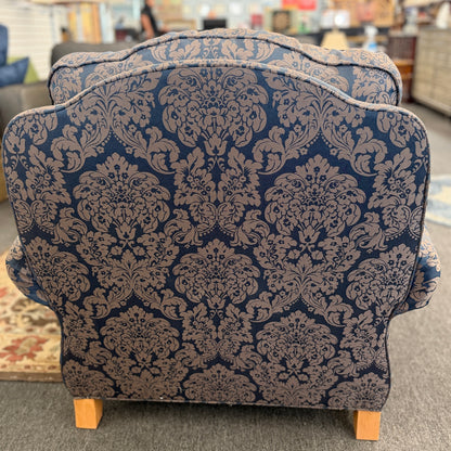 A traditional plush armchair featuring rolled arms and upholstered in a dark blue and brown damask pattern on a wooden frame.