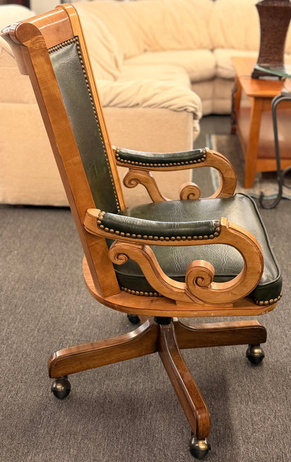 A traditional executive-style office desk chair with polished wood frame, deep forest green faux leather upholstery, brass nailhead trim, and a wheeled swivel base.