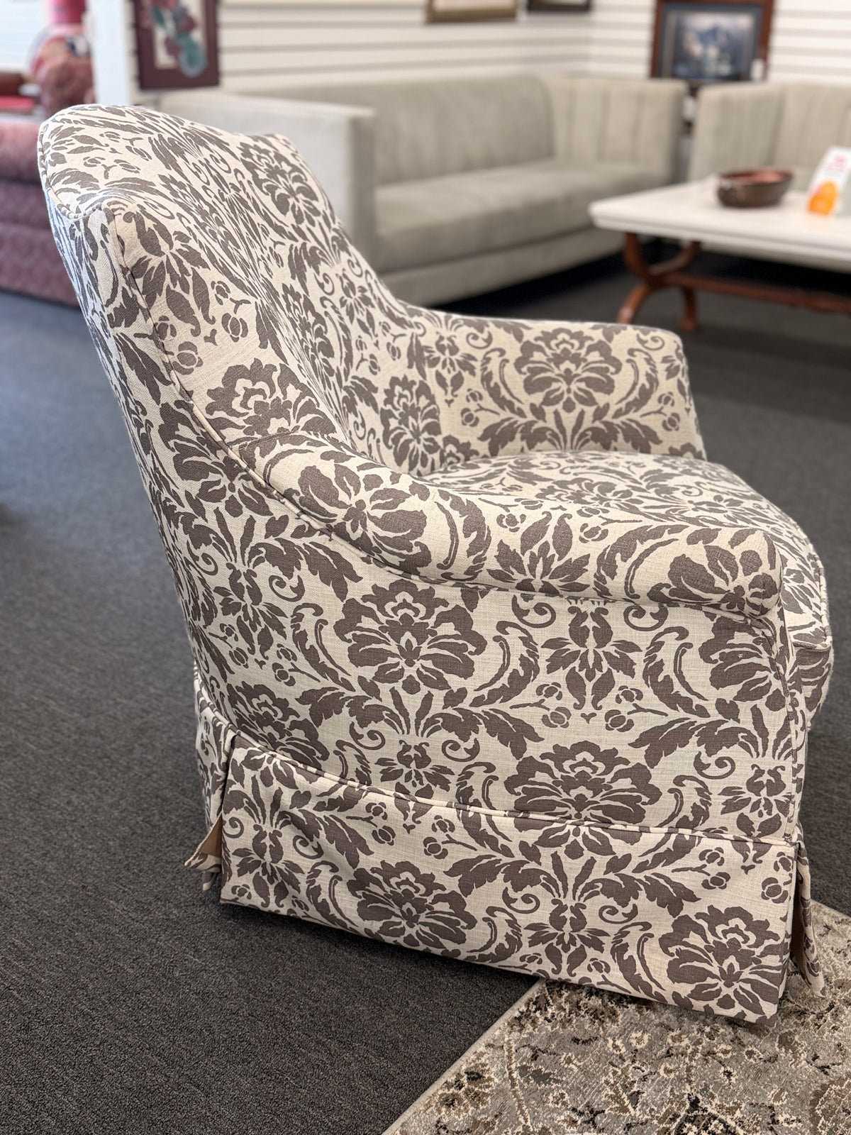 A traditional swivel armchair upholstered in a bold gray-and-cream floral damask pattern featuring a tailored base skirt in good condition.