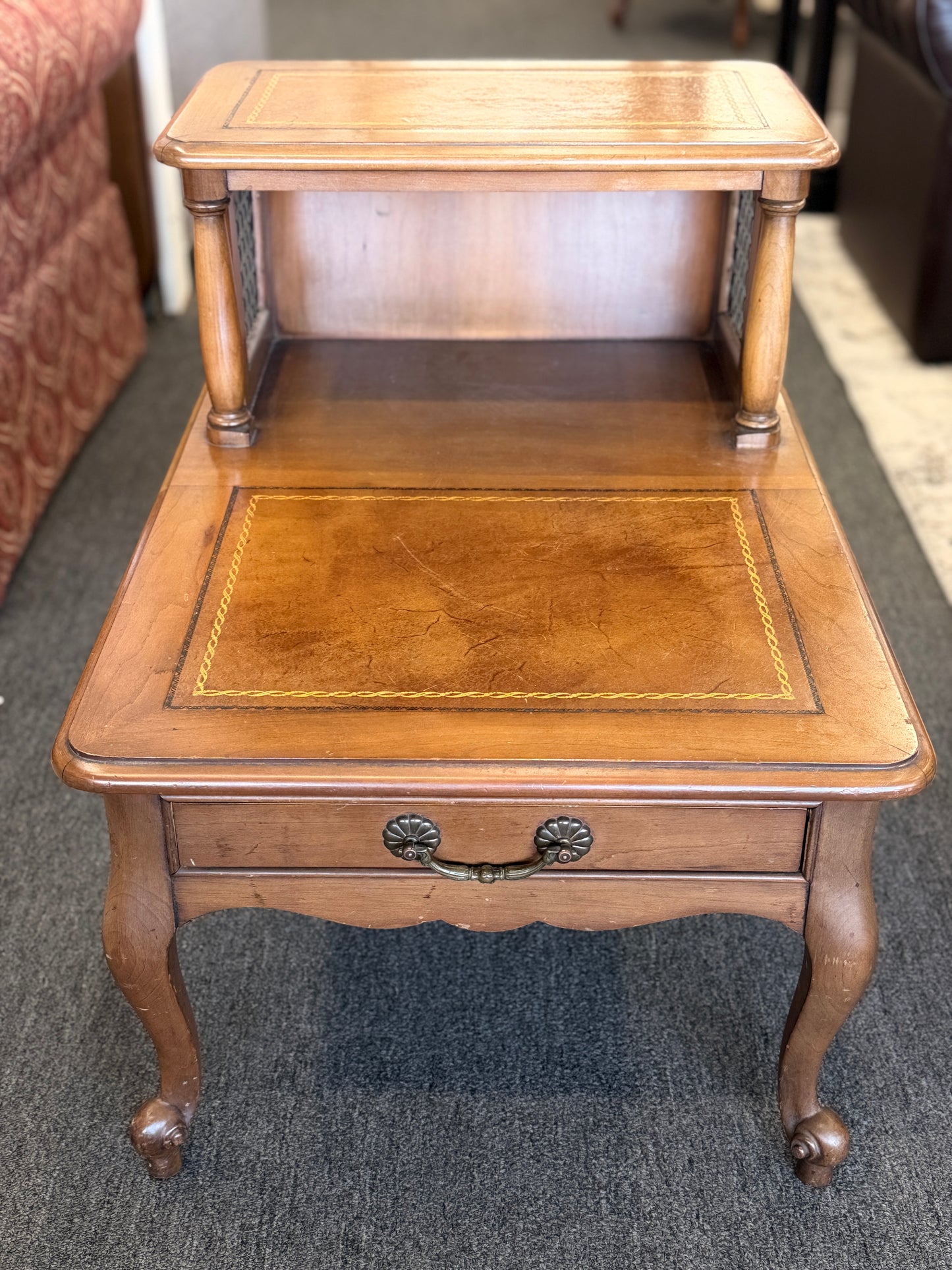 A wooden two-tiered step end table featuring decorative inlay with a gold border pattern, a single bottom drawer with ornate metal pulls, and cabriole legs.