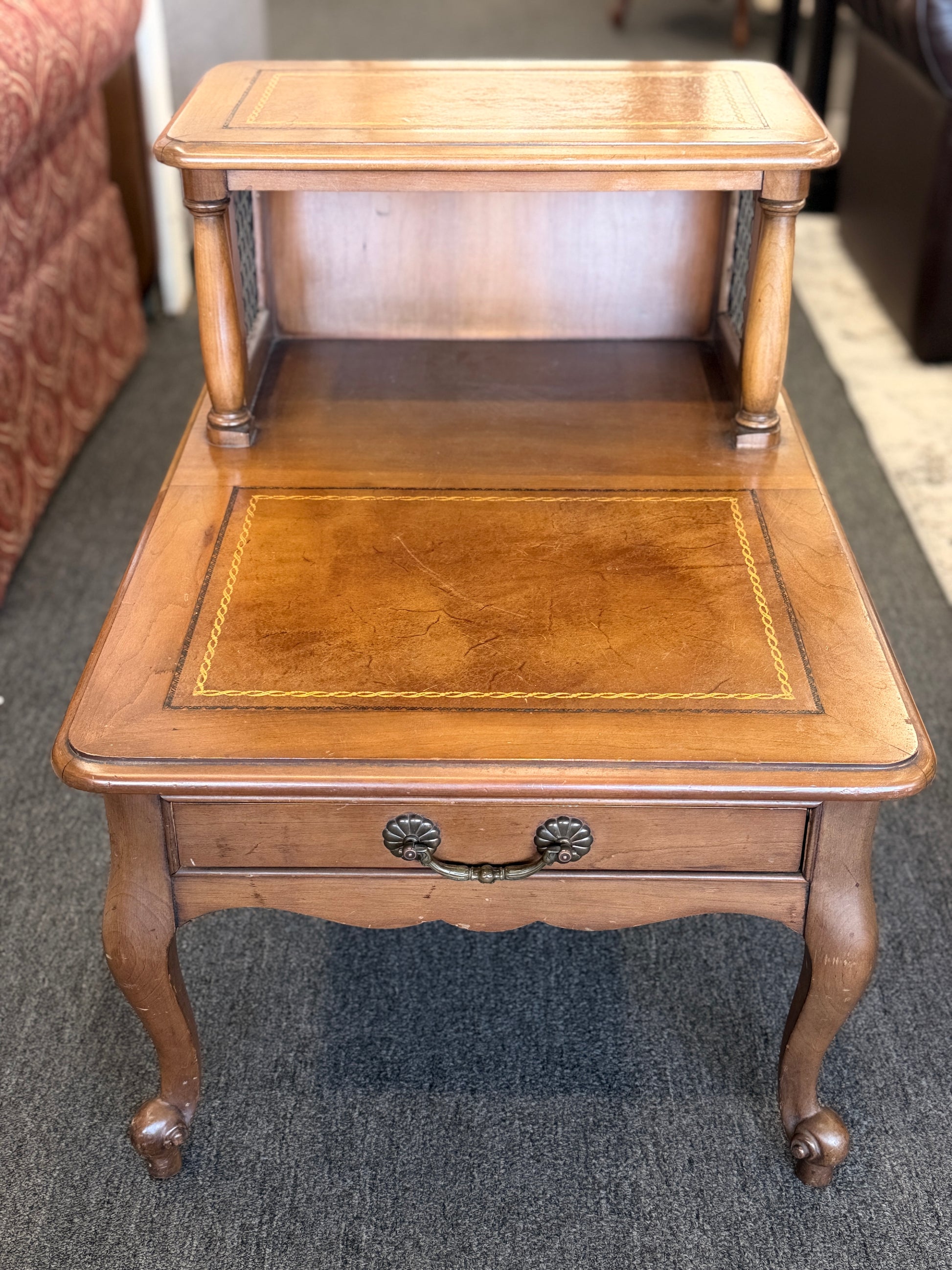 A wooden two-tiered step end table featuring decorative inlay with a gold border pattern, a single bottom drawer with ornate metal pulls, and cabriole legs.