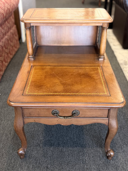 A wooden two-tiered step end table featuring decorative inlay with a gold border pattern, a single bottom drawer with ornate metal pulls, and cabriole legs.