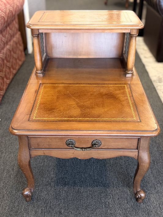 A wooden two-tiered step end table featuring decorative inlay with a gold border pattern, a single bottom drawer with ornate metal pulls, and cabriole legs.