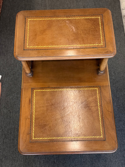 A wooden two-tiered step end table featuring decorative inlay with a gold border pattern, a single bottom drawer with ornate metal pulls, and cabriole legs.