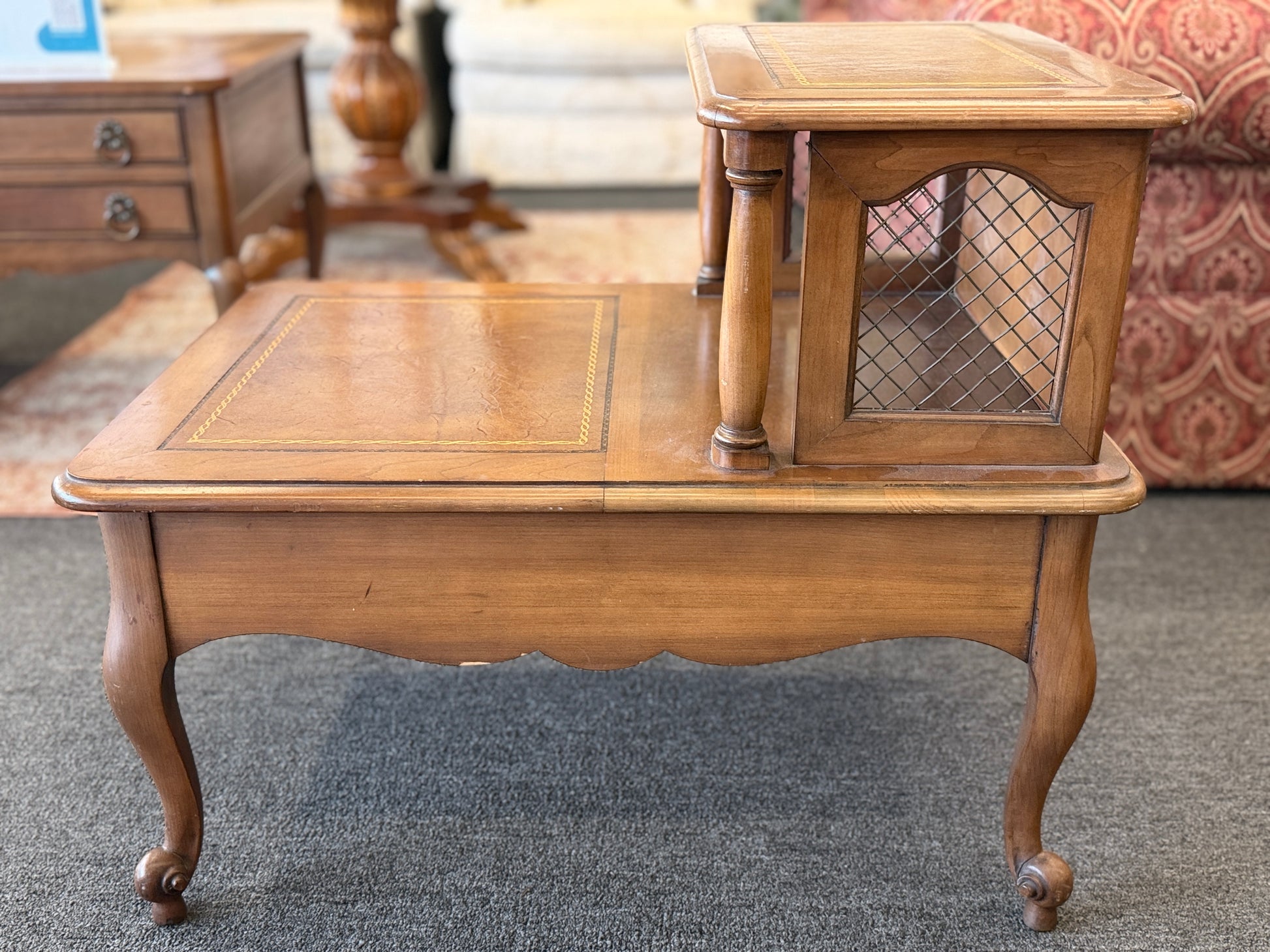 A wooden two-tiered step end table featuring decorative inlay with a gold border pattern, a single bottom drawer with ornate metal pulls, and cabriole legs.