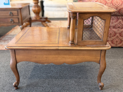 A wooden two-tiered step end table featuring decorative inlay with a gold border pattern, a single bottom drawer with ornate metal pulls, and cabriole legs.