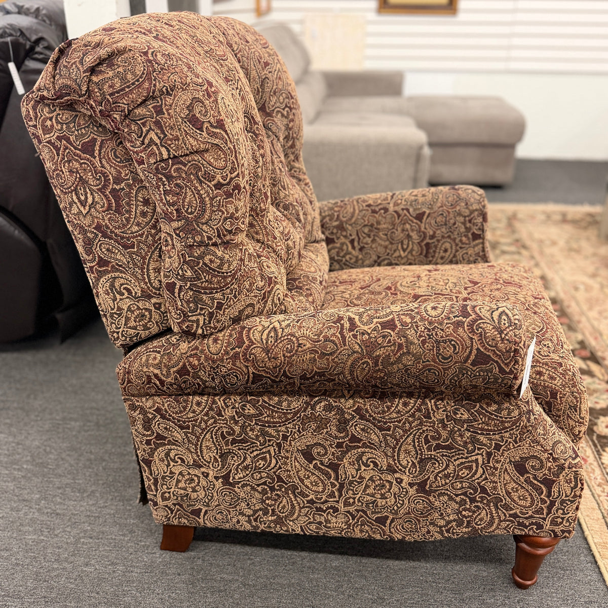 A traditional-style recliner armchair with rich brown and tan paisley upholstery, a tufted high backrest, rolled arms, and turned wooden legs in excellent condition.