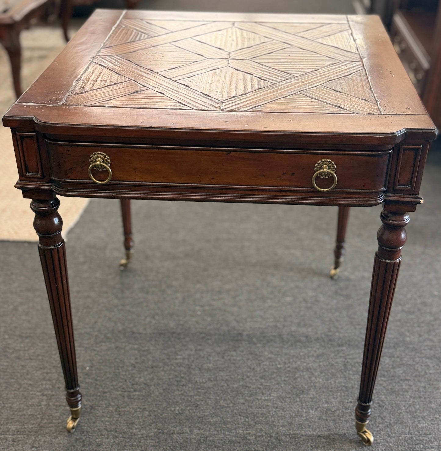 A classic 26-inch square wood end table featuring a geometric parquet inlay top, one drawer with a brass ring pull, and fluted legs on small caster wheels.