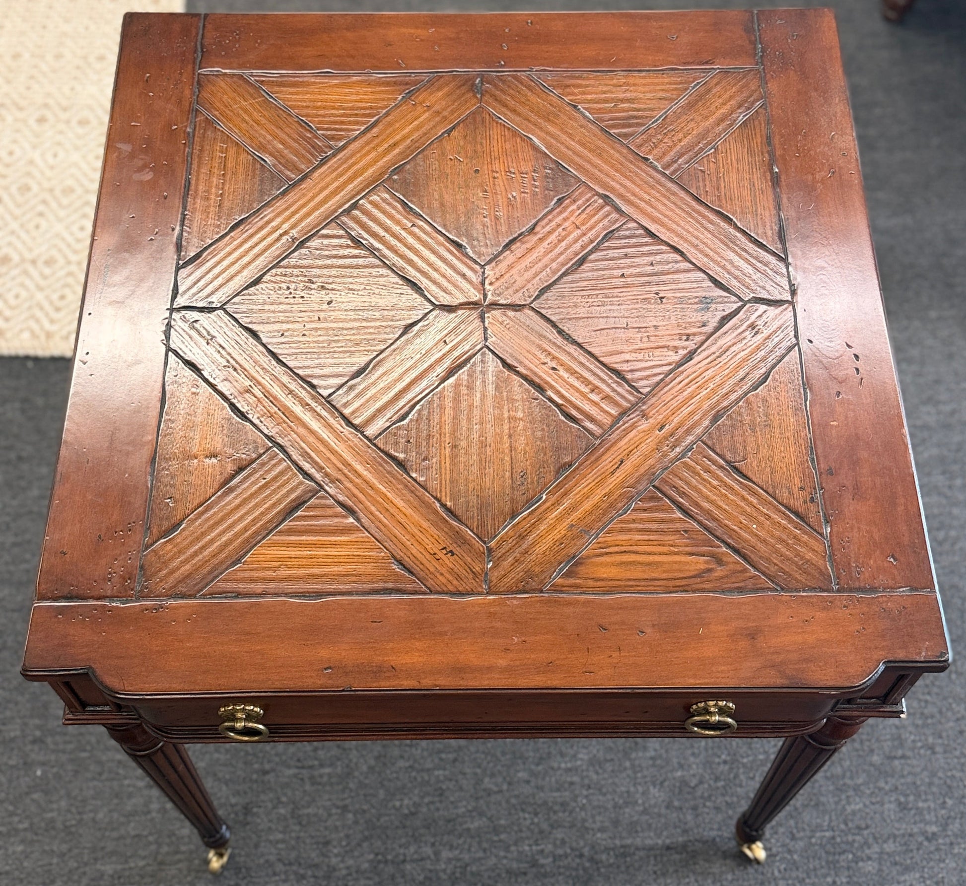 A classic 26-inch square wood end table featuring a geometric parquet inlay top, one drawer with a brass ring pull, and fluted legs on small caster wheels.
