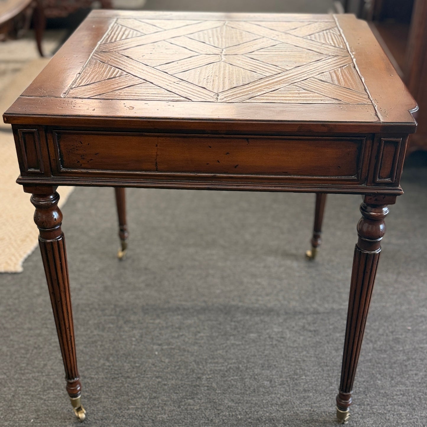 A classic 26-inch square wood end table featuring a geometric parquet inlay top, one drawer with a brass ring pull, and fluted legs on small caster wheels.
