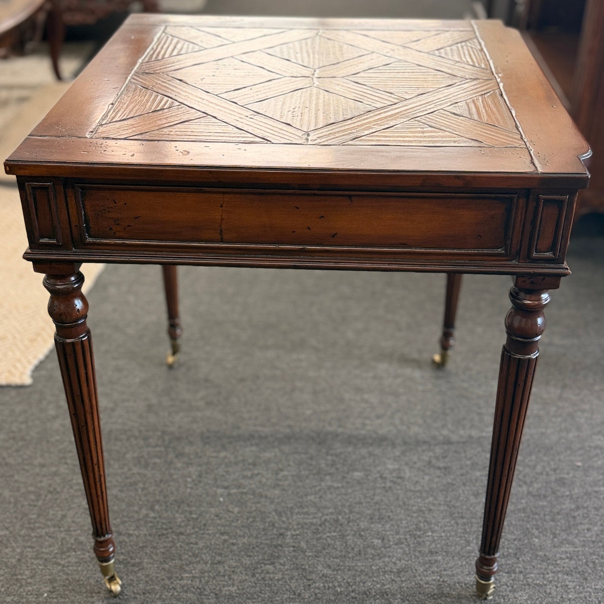 A classic 26-inch square wood end table featuring a geometric parquet inlay top, one drawer with a brass ring pull, and fluted legs on small caster wheels.