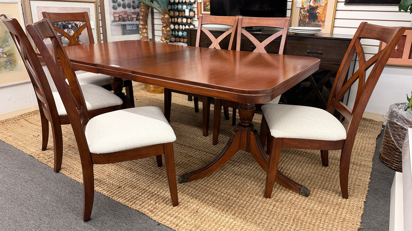 A traditional rectangular wooden dining table with an extendable leaf, twin turned pedestal bases, and six matching X-back dining chairs with light padded seats.