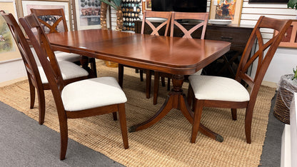 A traditional rectangular wooden dining table with an extendable leaf, twin turned pedestal bases, and six matching X-back dining chairs with light padded seats.