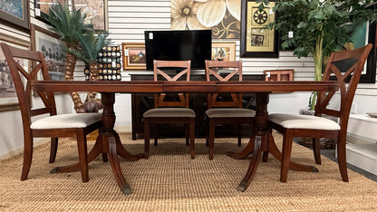 A traditional rectangular wooden dining table with an extendable leaf, twin turned pedestal bases, and six matching X-back dining chairs with light padded seats.