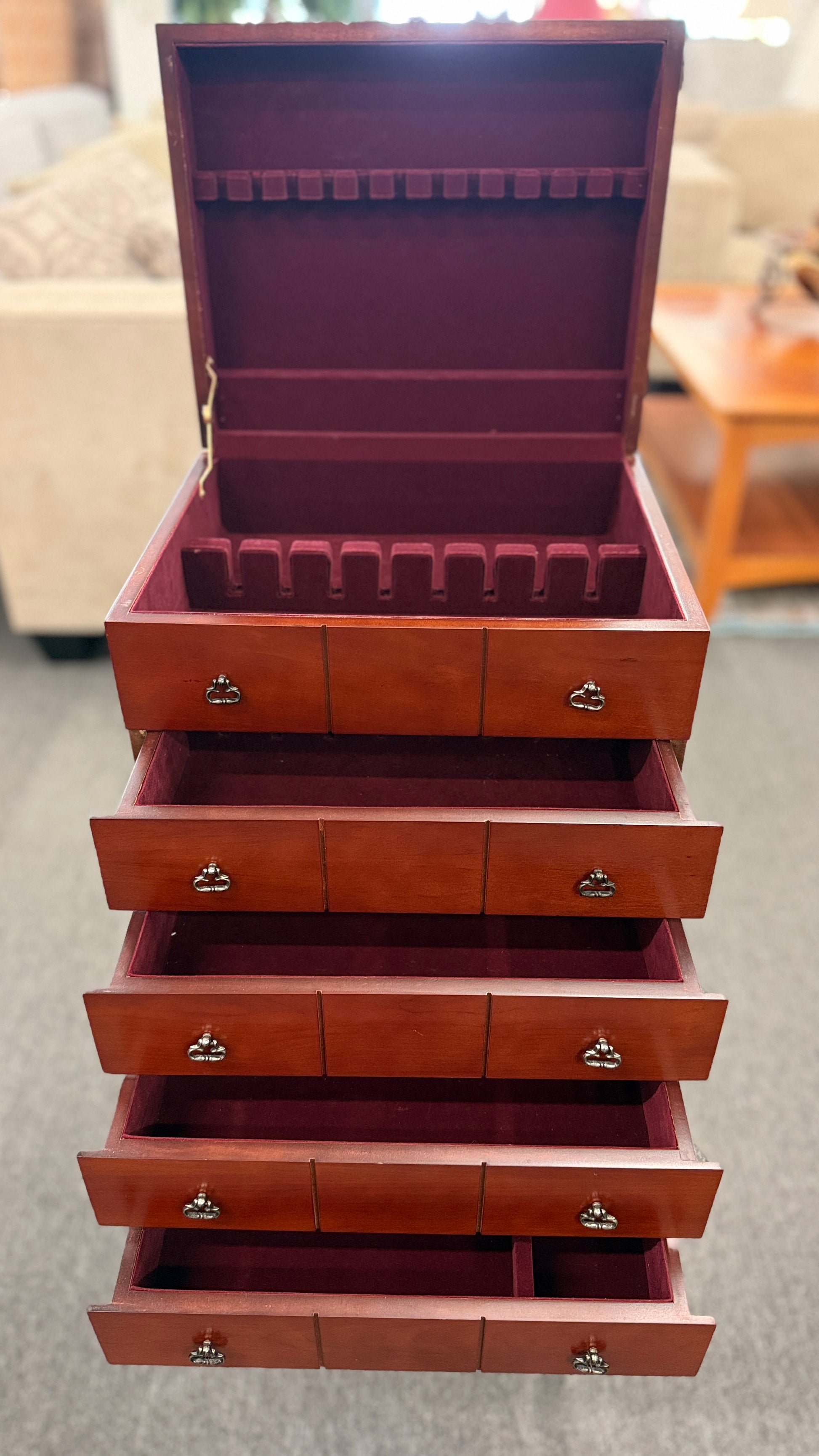 A tall, upright wooden jewelry or silverware chest shown open, revealing a ring-roll top compartment, five stacked drawers, burgundy fabric lining, and metal pulls.