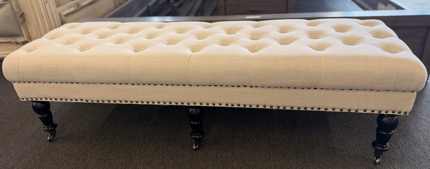 A long rectangular bedroom bench upholstered in cream linen with deep button tufting, antique bronze nailhead trim, and six black turned legs on casters in good condition.