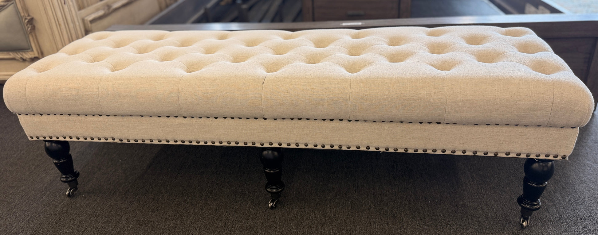 A long rectangular bedroom bench upholstered in cream linen with deep button tufting, antique bronze nailhead trim, and six black turned legs on casters in good condition.