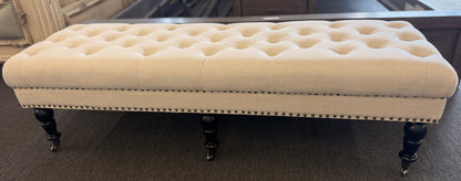 A long rectangular bedroom bench upholstered in cream linen with deep button tufting, antique bronze nailhead trim, and six black turned legs on casters in good condition.