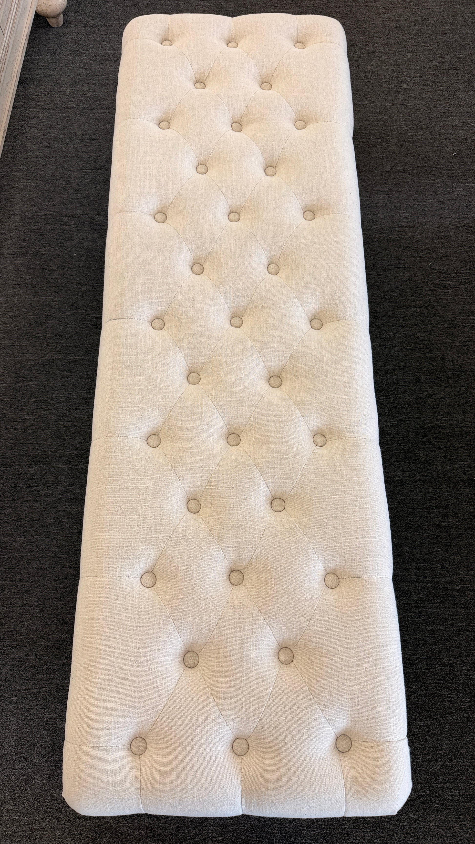 A long rectangular bedroom bench upholstered in cream linen with deep button tufting, antique bronze nailhead trim, and six black turned legs on casters in good condition.