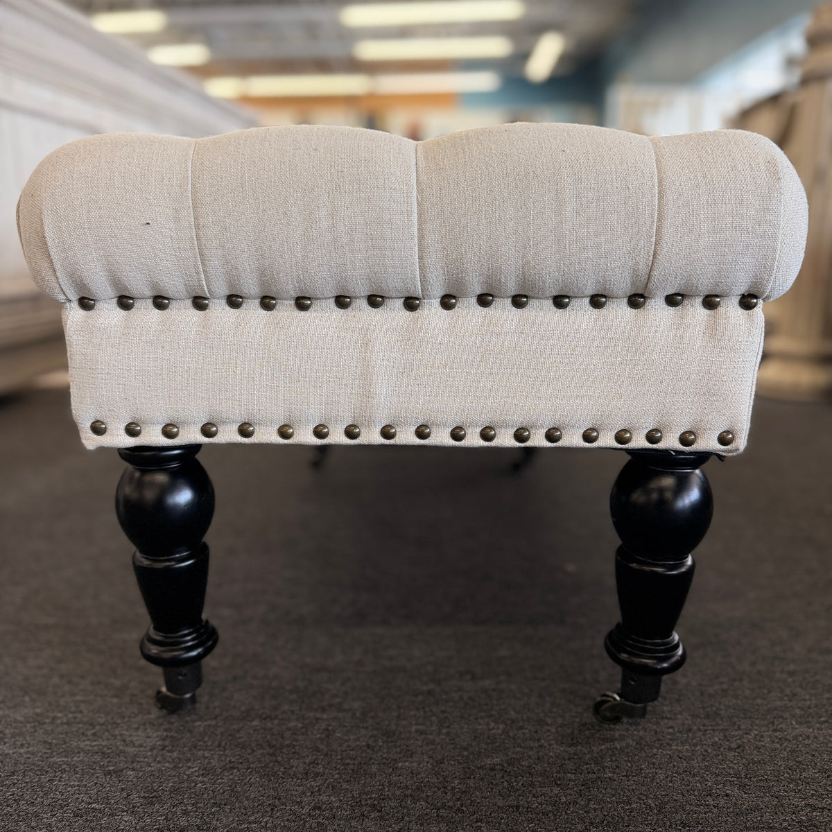 A long rectangular bedroom bench upholstered in cream linen with deep button tufting, antique bronze nailhead trim, and six black turned legs on casters in good condition.