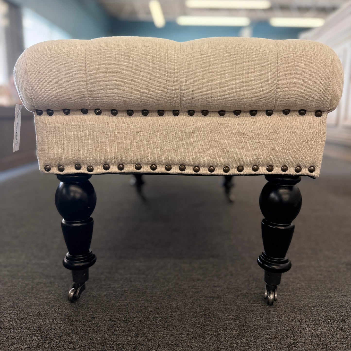 A long rectangular bedroom bench upholstered in cream linen with deep button tufting, antique bronze nailhead trim, and six black turned legs on casters in good condition.
